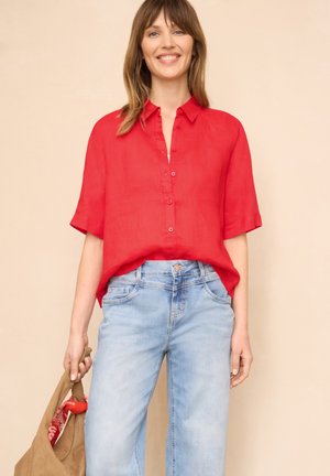 Lächelnde Frau, die ein locker sitzendes rotes Hemd und hellblaue Jeans trägt, und eine beige Tasche mit einem roten Schal darin hält.