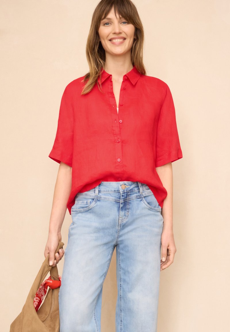 Femme souriante portant une chemise rouge ample à boutons et un jean bleu clair, tenant un sac beige avec une écharpe rouge à l’intérieur.