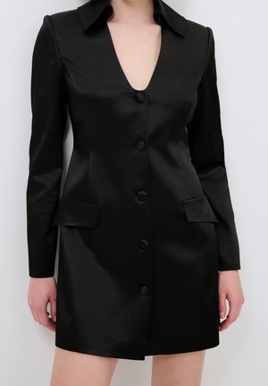 Nainen pukeutuneena istuvaan mustaan satiiniseen bleiserimekkoon, jossa on pitkät hihat, syvä V-pääntie, etunapit ja läppätaskut, seisomassa yksiväristä taustaa vasten.