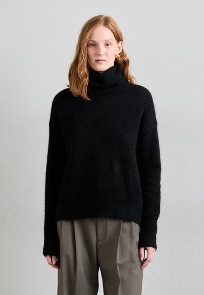 Czarny sweter typu golf w miękkim, puszystym materiale. Luźny krój z długimi rękawami, noszony z jasnoszarymi, wysokogatunkowymi spodniami.