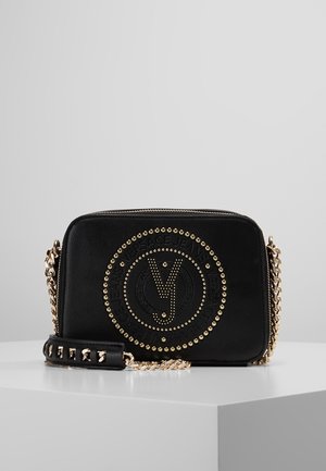 Sac bandoulière en cuir noir avec des clous dorés formant le logo circulaire Versace Jeans et une sangle chaîne dorée, posé sur une surface blanche.