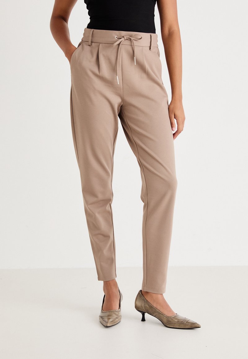 Donna che indossa pantaloni beige su misura con vita regolabile a coulisse e scarpe metalliche con tacco basso e punta affilata, in piedi davanti a uno sfondo semplice.