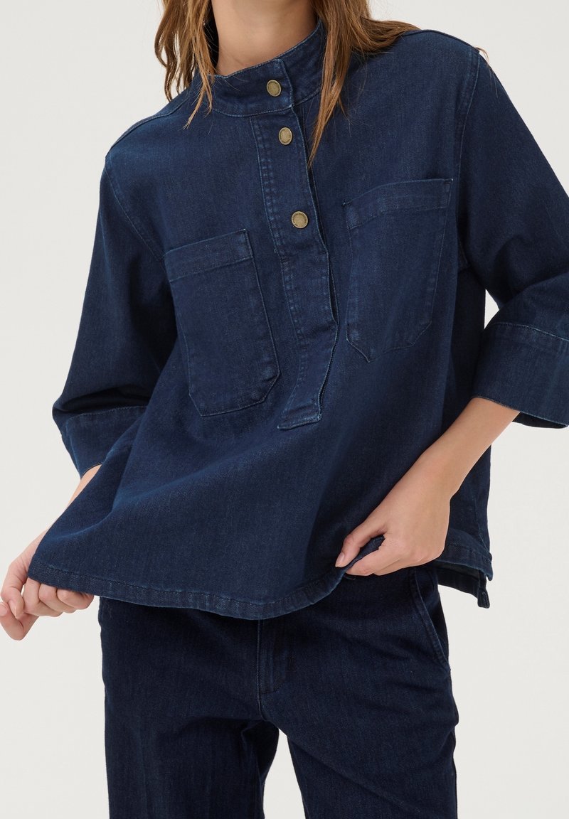 Femme portant une chemise en denim bleu foncé avec trois boutons en laiton et deux poches poitrine, assortie à un pantalon en denim bleu foncé.