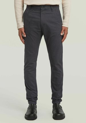 Persona che indossa pantaloni grigio scuro slim-fit con sottili righe e scarpe di pelle nere, in piedi contro uno sfondo chiaro.