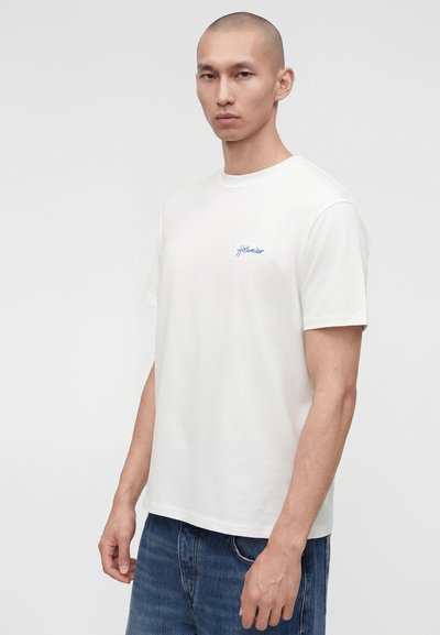 Άνδρας με ξυρισμένο κεφάλι που φοράει λευκό κοντομάνικο μπλουζάκι με μικρό μπλε κείμενο στο στήθος, συνδυασμένο με μπλε τζιν, μπροστά από απλό φόντο.