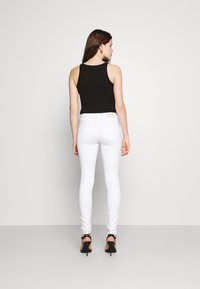 Top preto de alças finas, combinado com calças brancas justas e sapatos de salto alto pretos, visto de costas contra um fundo simples.