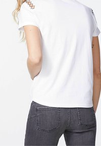 Bílý bavlněný tričko s kulatým výstřihem a dekorativním stříbrným kováním na rameni, v kombinaci se šedými džíny s dvěma zadními kapsami.