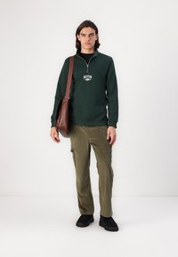 Grønn zip-up hettegenser med "BOSTON" trykk, kombinert med olivenfargede corduroy cargo bukser og svarte slip-on sko; brun skulderveske.