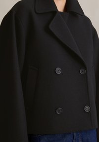 Veste noire à double boutonnage en tissu texturé, dotée de grands boutons noirs, d'un col à revers et de poches latérales. Design court.