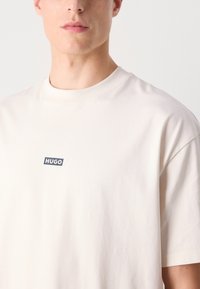 Cremfarbendes Baumwoll-T-Shirt mit Rundhalsausschnitt und kurzen Ärmeln, versehen mit einem blauen rechteckigen Logo mit der Aufschrift "HUGO" auf der Vorderseite.