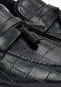 Zwarte leren instappers met een gestructureerd krokodillenpatroon, voorzien van een decoratieve strik en kwastdetail aan de voorkant.