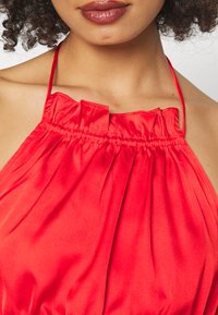 Top halter vermelho feito de cetim suave com decote franzido e alças. Apresenta um tecido solto e fluido que cria uma silhueta suave.