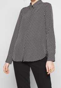 Blusa a maniche lunghe con pois bianchi e neri, colletto, chiusura a bottoni e maniche con orlo, indossata sopra pantaloni neri. Tessuto dalla superficie liscia.