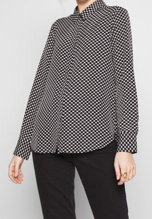 Blusa a maniche lunghe con pois bianchi e neri, colletto, chiusura a bottoni e maniche con orlo, indossata sopra pantaloni neri. Tessuto dalla superficie liscia.