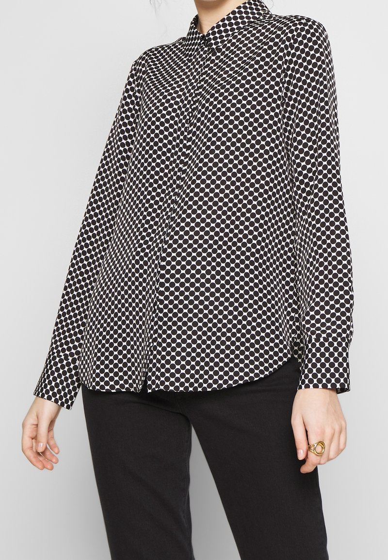 Blusa a maniche lunghe con pois bianchi e neri, colletto, chiusura a bottoni e maniche con orlo, indossata sopra pantaloni neri. Tessuto dalla superficie liscia.