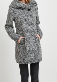 Donna che indossa un cappotto di lana grigia con texture, colletto ampio, chiusura a bottoni e mani nelle tasche anteriori, abbinato a pantaloni neri.