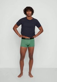 Mlad muškarac s kovrčavom kosom u crnoj majici i zelenim Levi's boksericama, stoji bos naslonjen na običnu sivu pozadinu.