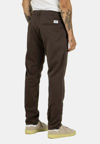 Braune Baumwollhosen mit entspannter Passform, ausgestattet mit einer Gesäßtasche und einem kleinen Markenetikett. Getragen mit beigen Sneakers.