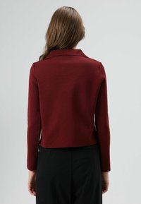 Blazer bordeaux avec un col structuré et des manches longues, en tissu texturé et de coupe courte, associé à un pantalon noir.