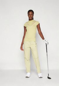 Mulher em vestuário de golfe amarelo claro segurando um taco de golfe, usando luva branca e ténis brancos, em pé contra um fundo simples.