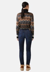 Pletený svetr s geometrickým vzorem v černé, hnědé a bílé; spárován s tmavě modrými džíny; model má na sobě béžové boty s kožešinovým lemem.