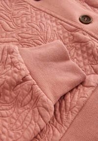 Manșeta cu quilt roz a unei jachete, cu margine zimțată și detalii florale brodate, arătând o parte dintr-un nasture maro pe țesătură.