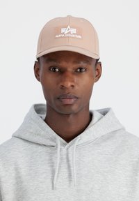 Căciulă bej cu vizieră curbată și patch alb cu logo-ul "Alpha Industries". Pur tată cu un hanorac gri deschis, dotat cu șnur pentru ajustare la gât.