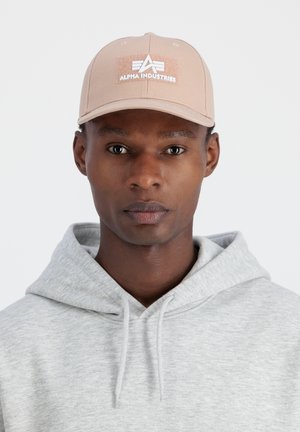 Bézs sapka ívelt karimával és fehér "Alpha Industries" logóval. Világosszürke kapucnis pulóverrel viselve, amelyen húzózsinór található a nyaknál.