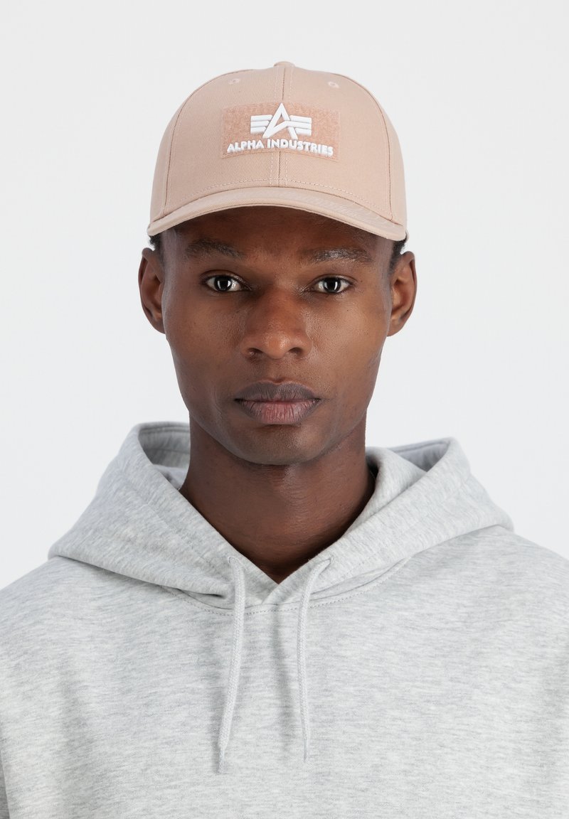 Căciulă bej cu vizieră curbată și patch alb cu logo-ul "Alpha Industries". Pur tată cu un hanorac gri deschis, dotat cu șnur pentru ajustare la gât.