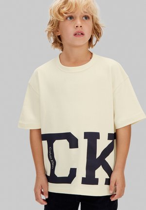 Copil blond purtând un tricou crem supradimensionat cu litere mari negre „CK” și textul „Calvin Klein”, stând pe un fundal gri.