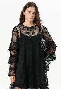 Svart blondekjole med lange, volangermede ermer, en gjennomsiktig bodice og en flytende silhuett; intrikat blomsterdesign gjennom hele.
