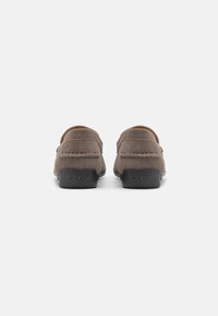 Mocasines de ante marrón con una suela de goma negra baja, que presentan un pespunte decorativo y un logo sutil en el talón.