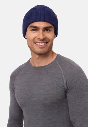 Navy knitted beanie with a ribbed texture, featuring a small brown leather label. Worn with a fitted gray long-sleeve shirt.