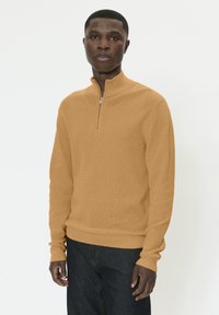 Homme portant un pull à demi-fermeture éclair de couleur beige et un jean foncé, debout devant un fond blanc uni, regardant droit devant lui.