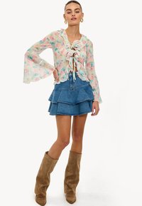 Blouse légère et transparente à motif floral avec un col volanté et des manches cloche, associée à une mini-jupe en jean à volants et des bottes hauteur genou couleur sable.