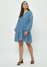 Blau gemustertes Kleid mit langen Ärmeln, V-Ausschnitt und gestuftem Saum. Hergestellt aus leichtem Material mit geometrischem Druck. Braune Sandalen mit Blockabsatz.
