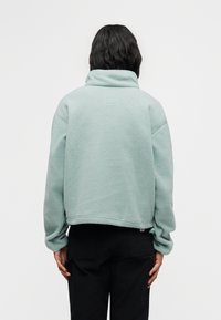 Άτομο που φαίνεται από πίσω φοράει ανοιχτό μπλε μπουφάν φλίς με ψηλό γιακά και μαύρο παντελόνι στο φόντο απλής λευκής επιφάνειας.