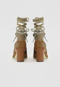 Brown heeled shoes with a smooth texture, featuring a metallic gold accent at the ankle and crisscross laces in olive green.