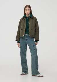 Khaki bomber jacket z zielonym swetrem pod spodem, w połączeniu z szerokimi niebieskimi dżinsami. Posiada kieszenie i zamek błyskawiczny.