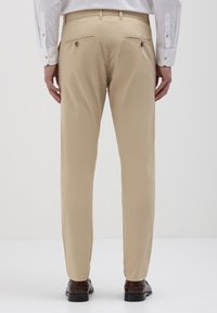 Pantaloni khaki realizzati in un tessuto liscio, dotati di due tasche posteriori con chiusura a bottone e un design a gamba dritta. Indossati con scarpe scure.