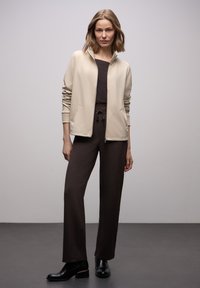 Giacca beige con cerniera sopra una maglia marrone scuro e pantaloni coordinati, realizzati in tessuto morbido, con una vestibilità comoda e tasche. Stivaletti neri alla caviglia.