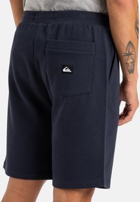 Pantalones cortos de algodón azul marino con cintura elástica, bolsillo trasero y un parche con logo en la parte posterior, que presentan una textura suave y un diseño sencillo.