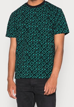 Mann trägt ein türkisfarbenes T-Shirt mit einem schwarzen geometrischen Labyrinthmuster und schwarze Hosen, steht mit entspannten Armen an den Seiten.