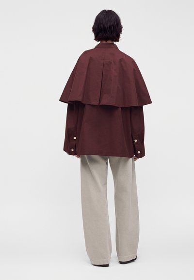 Personne vue de dos portant une veste bordeaux ample avec un détail de cape dans le dos et un pantalon large beige sur un fond uni.
