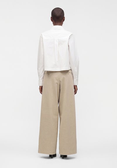 Personne aux cheveux courts portant une chemise blanche à manches longues et un pantalon beige à jambes larges, debout de dos sur un fond uni.