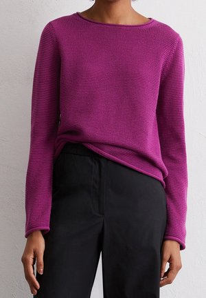Personne portant un pull à manches longues texturé de couleur magenta légèrement rentré dans un pantalon noir taille haute, sur un fond uni.