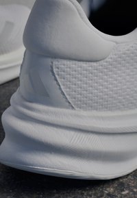 Zapatilla deportiva blanca con tejido texturizado y superposiciones suaves. Cuenta con un cuello de talón acolchado y un sutil detalle del logo en el costado.