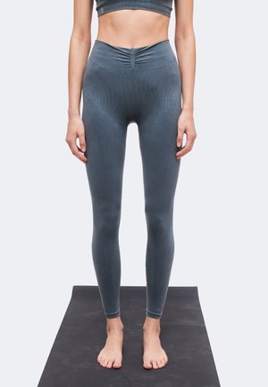 Persona de pie descalza sobre una esterilla de yoga negra, llevando mallas sin costuras de tiro alto de color gris oscuro y un sujetador deportivo a juego.