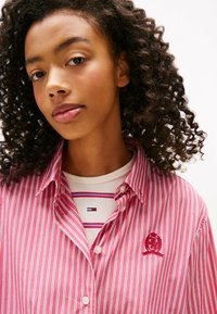 Czerwono-biała koszula w paski z kołnierzykiem zapinanym na guziki, z haftowanym logo na piersi, noszona na koszuli w paski.