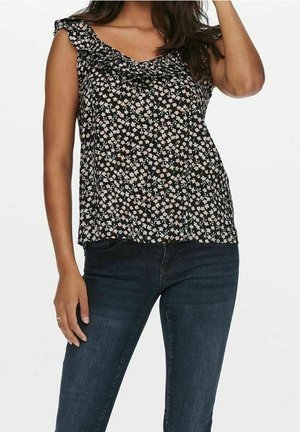 Woman wearing sleeveless black floral blouse with ruffle neckline and dark blue fitted jeans against white background.